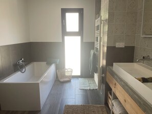 Ett modernt badrum med badkar, dusch och handfat med tillhörande skåp. Det står en korg på golvet och en handduk hänger på väggen.