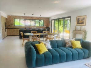 Una sala de estar moderna con un sofá color turquesa, una zona de comedor con sillas de madera y una cocina con pared de ladrillo y ventanas amplias.