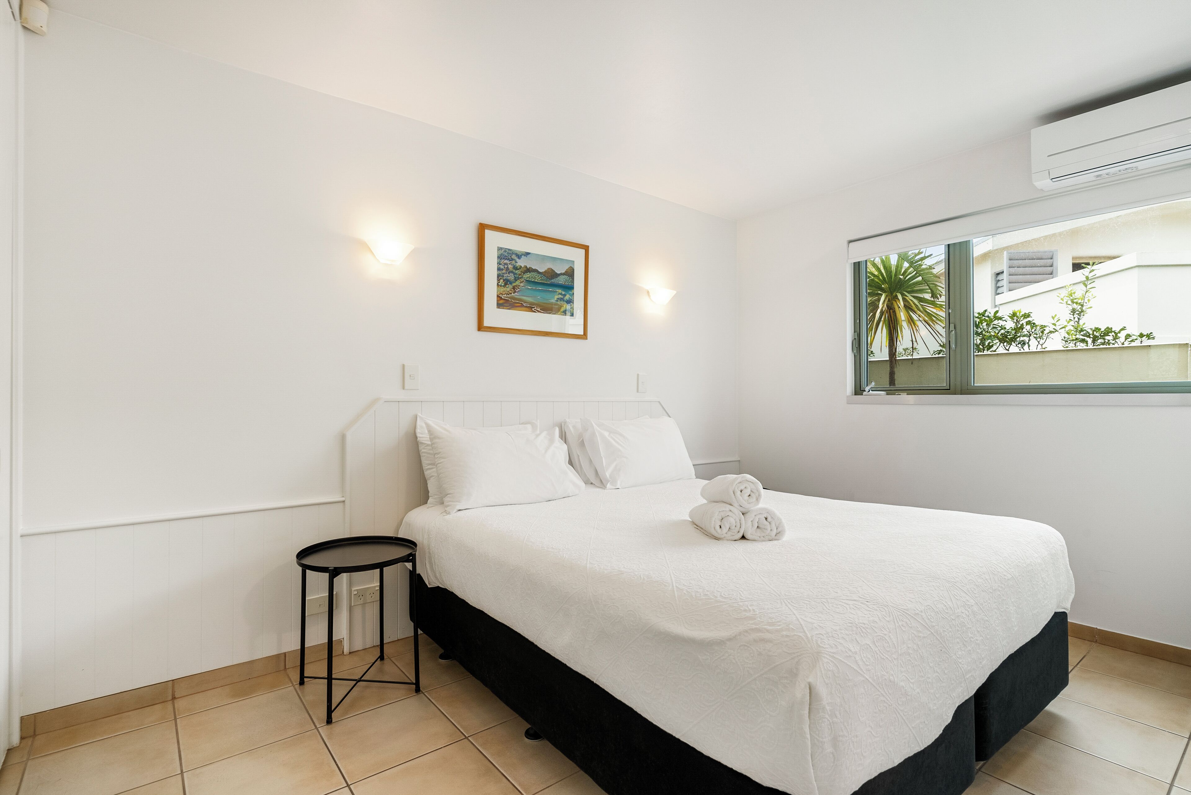Een netjes opgemaakt bed met witte lakens en een klein nachtkastje, een ingelijste foto aan de muur, en een raam met uitzicht op groen.