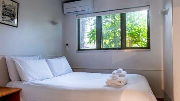 A hotel room with a bed, white linens, a window with a view of greenery, an air conditioning unit, and a framed picture on the wall.