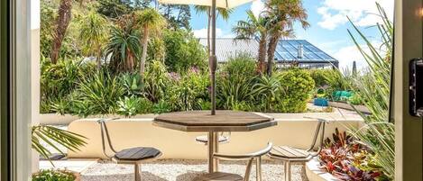 Une terrasse extérieure avec une table ronde et deux chaises, entourée de différentes plantes et d’un grand parasol.