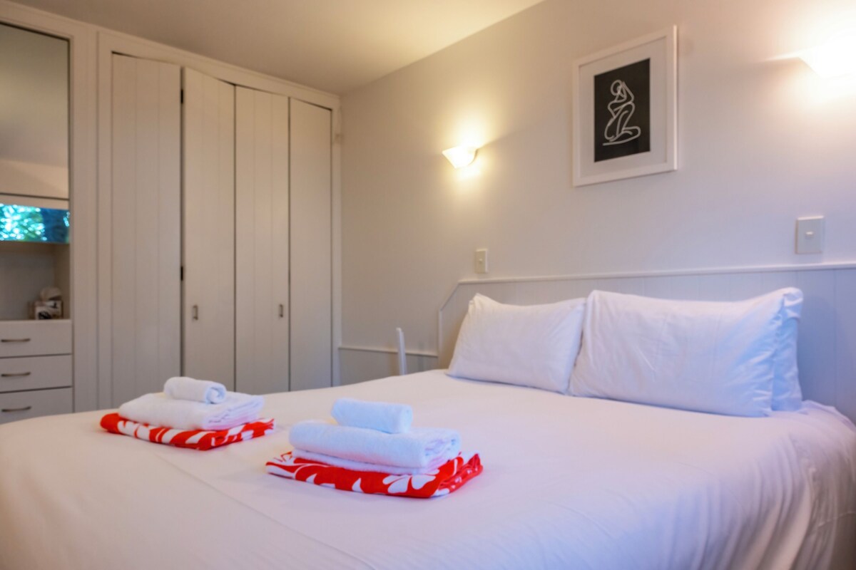 A hotel room with a bed, white pillows, a headboard, a wall-mounted light, a framed picture, and a wardrobe with a mirror.