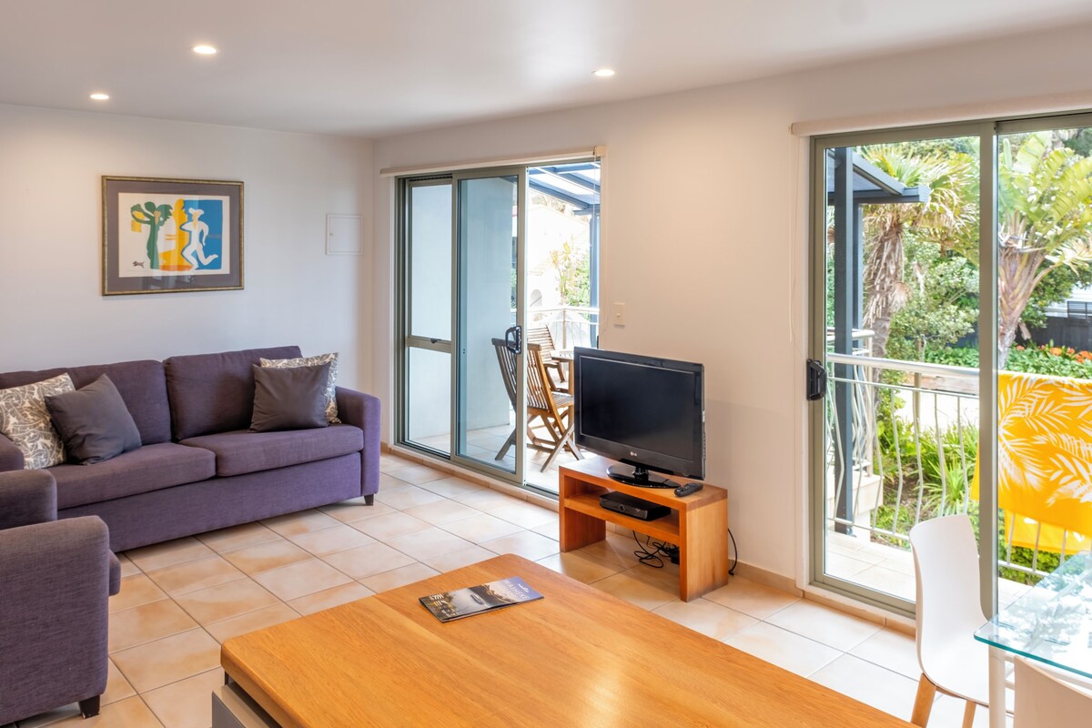 A living room with a sofa, a coffee table, a TV on a wooden stand, and a sliding glass door leading to a balcony.