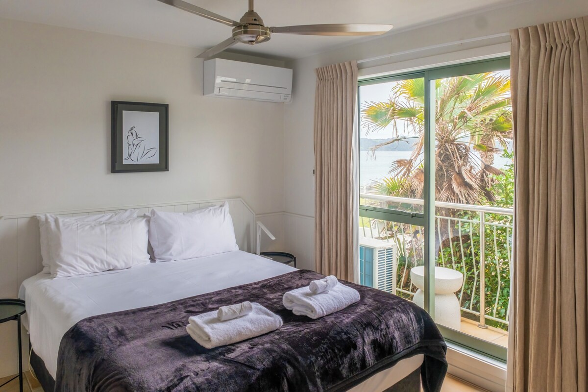 A hotel room with a bed, a ceiling fan, an air conditioning unit, a framed artwork, and a balcony with a view of palm trees and a body of water.