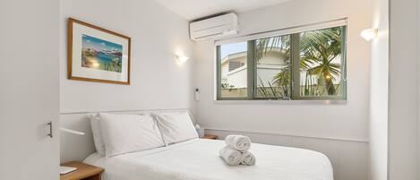 A neatly made bed with white linens, a framed picture on the wall, and a window with a view of palm trees.