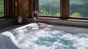 Un bain à remous, un verre de vin rouge et un livre posé sur un rebord de fenêtre, avec vue sur un paysage boisé.