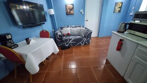 Un espacio habitable compacto con un sofá, una mesita con mantel blanco, un televisor montado en la pared, una cafetera y un extintor.