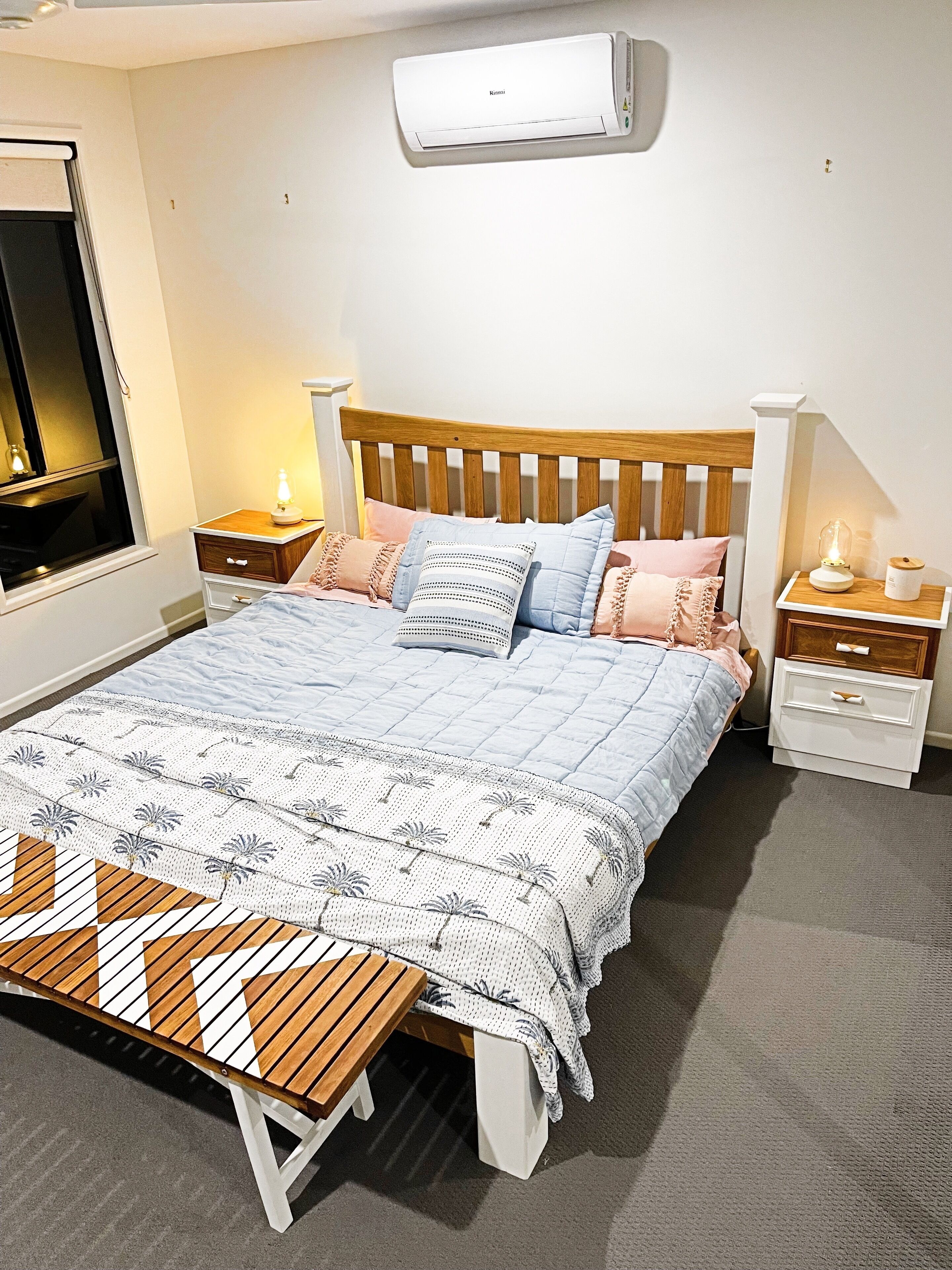 A bedroom with a bed, bedside tables, a nightstand, a lamp, a bench, and an air conditioning unit.