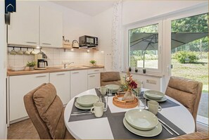 Een kleine keuken met witte kastjes, een magnetron en een koffiezetapparaat. Er is een eettafel met stoelen, een vaas met bloemen en een raam met uitzicht op groen.