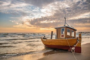 Un bateau en bois doté d’une cabine, amarré sur une plage de sable où les vagues se brisent, sous un ciel partiellement nuageux.