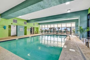 An indoor swimming pool with clear glass walls, a tiled floor, and a view of the outdoors. - Coastal Luxe | Meridian Six Oh Two (Myrtle Beach)