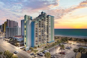 A high-rise hotel with multiple balconies, a parking lot, and a view of the beach at sunset.