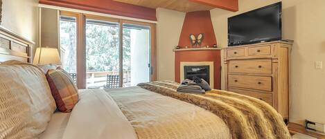 A bedroom with a large bed, a television, a fireplace, and a view of the outdoors.