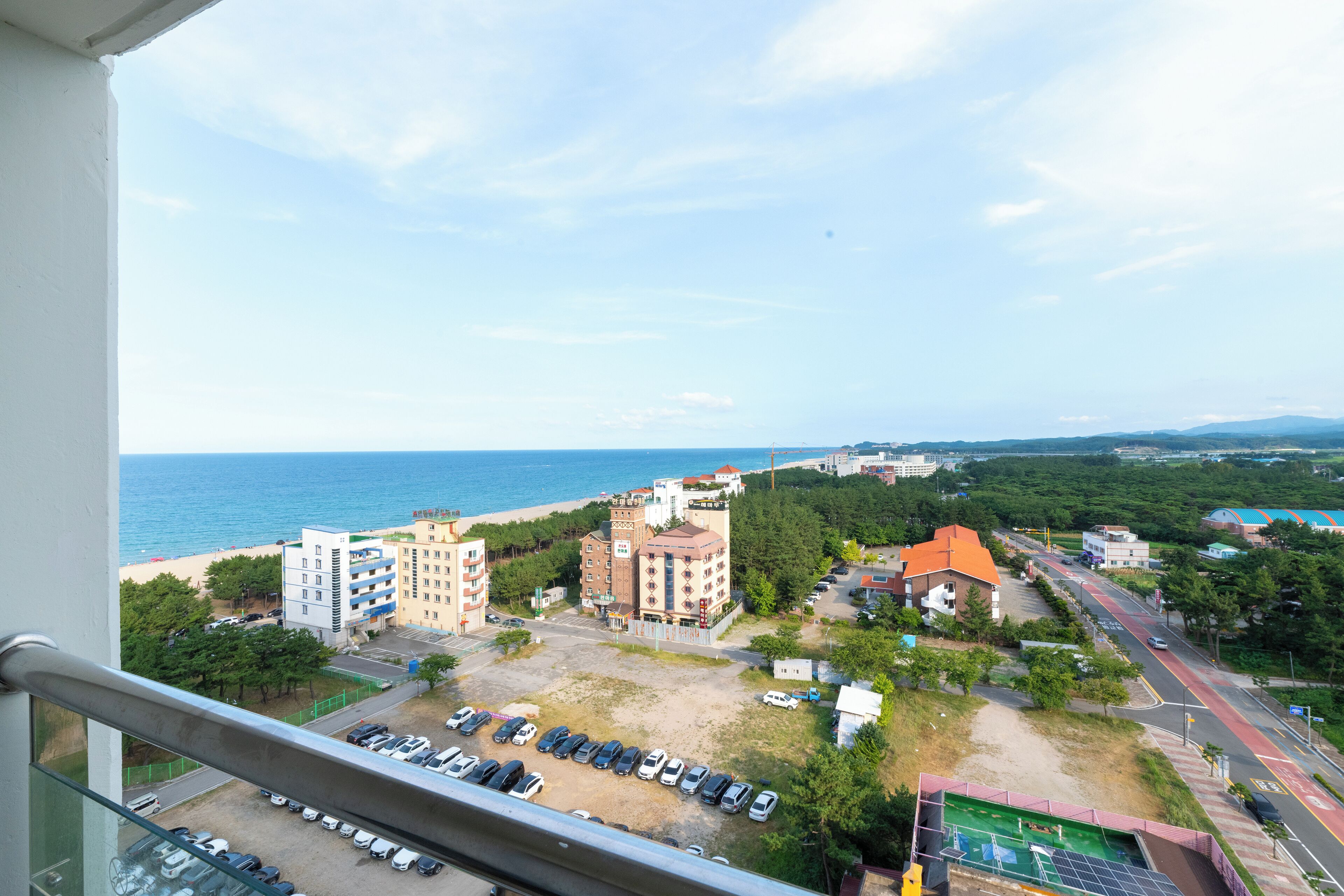Vista desde un balcón sobre un estacionamiento, una playa y edificios bajo un cielo despejado.