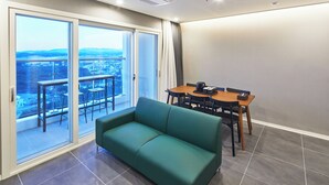 Un salon moderne avec un canapé vert, une table à manger avec des chaises et une grande porte-fenêtre coulissante donnant sur un balcon avec vue sur la ville.