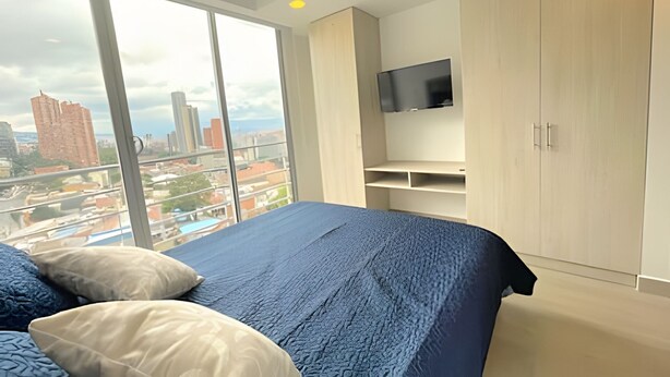 Un dormitorio con un ventanal grande, una cama con ropa de cama azul, un televisor montado en la pared y vistas a la ciudad.