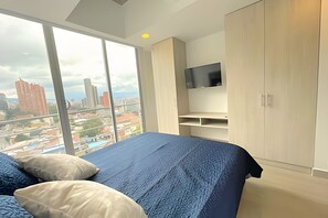 A bedroom with a large window, a bed with blue bedding, a TV mounted on the wall, and a city view. - Apartamento en Centro Internacional  (Bogotá)