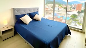 Un dormitorio moderno con una cama grande, una mesita de noche y vistas a un paisaje urbano a través de amplias ventanas.