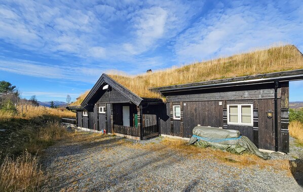 Ein rustikales Blockhaus mit Schotterauffahrt, umgeben von hohem Gras und wolkenlosem Himmel.