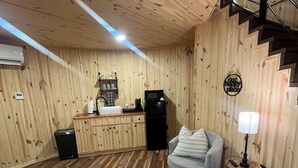 Tunnelmallinen mökin sisustus, jossa on puuseinät, puulattia, keittiöalue, jossa on pesuallas ja liesi, kaksi harmaata nojatuolia ja portaikko.