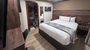 Une chambre d’hôtel comprenant un lit, un bureau, une chaise, une armoire, un miroir, une salle de bain avec un lavabo et des toilettes, et un luminaire au plafond.