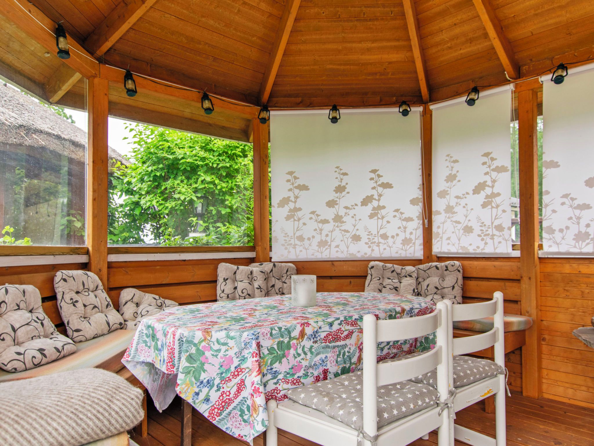 A covered outdoor seating area with a table and chairs, surrounded by wooden walls and windows with floral wallpaper.