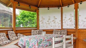 A covered outdoor seating area with a table and chairs, surrounded by wooden walls and windows with floral wallpaper.