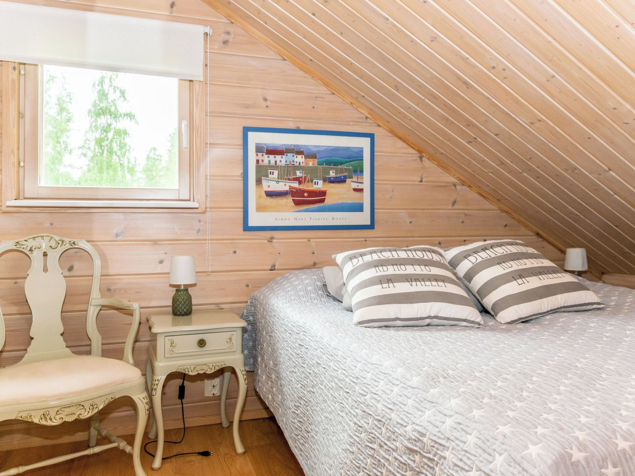 Ein gemütliches Schlafzimmer mit schrägem Dach, einem Bett mit gestreiften Kissen, einem Nachttisch mit Lampe, einem Sessel und einem gerahmten Bild von Booten.
