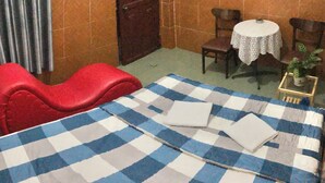 Ein Zimmer mit kariertem Bettbezug, einem roten Sessel, einem kleinen runden Tisch mit weißem Tischtuch und einer Topfpflanze.