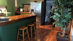 Une cuisine avec des meubles de couleur verte, un plan de travail en bois et un réfrigérateur en acier inoxydable. On y trouve trois tabourets et une plante en pot.