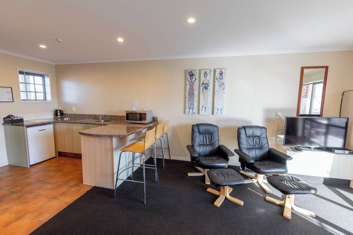 A modern kitchen with a bar area, two black leather chairs, a black ottoman, a flat-screen TV, and artwork on the wall.