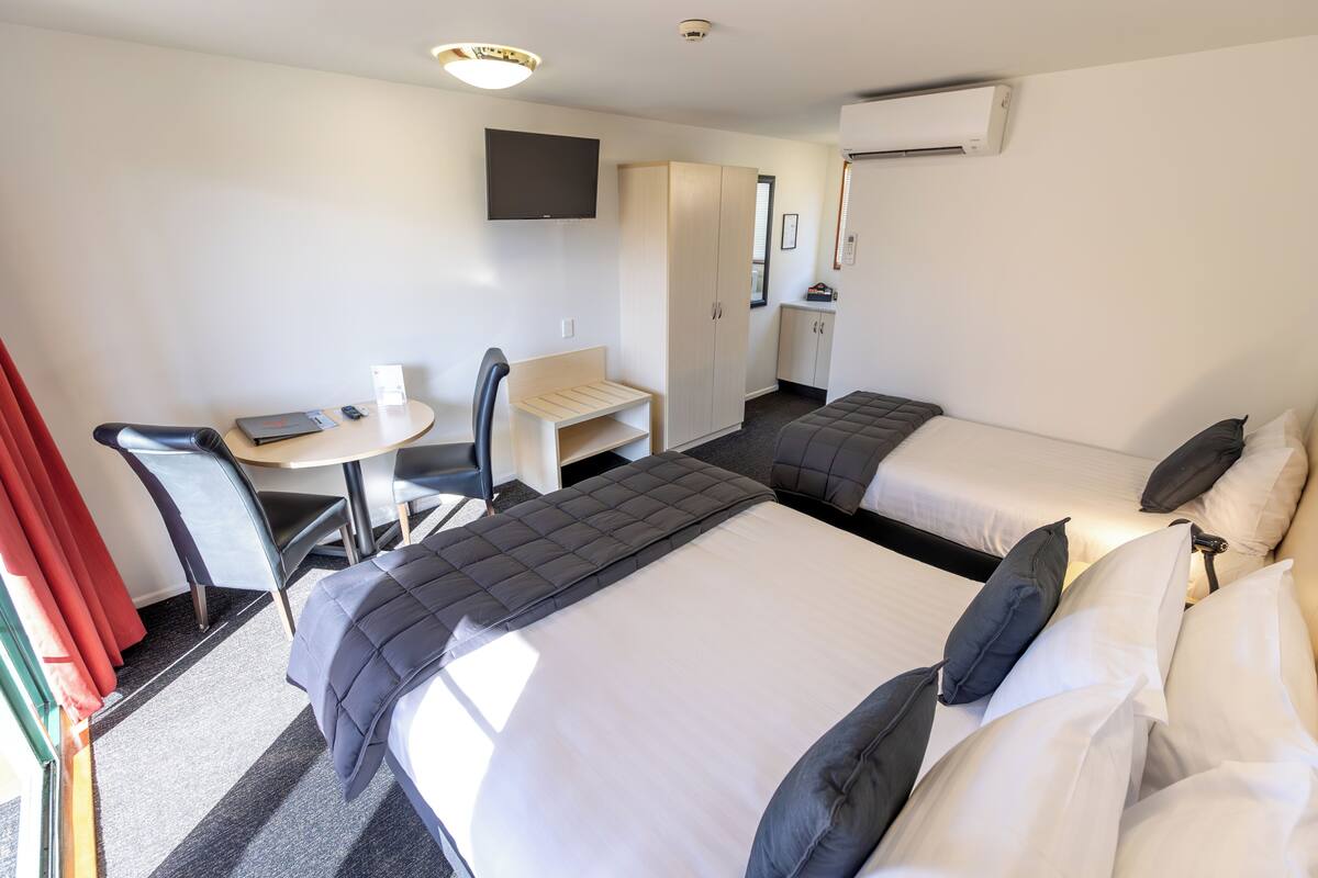 A hotel room with two beds, a desk, a chair, a TV, and an air conditioning unit.