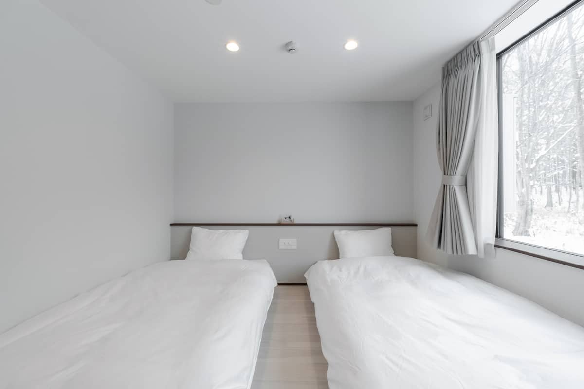 A modern, minimalist room with two single beds, a large window with curtains, and a ceiling with recessed lighting.
