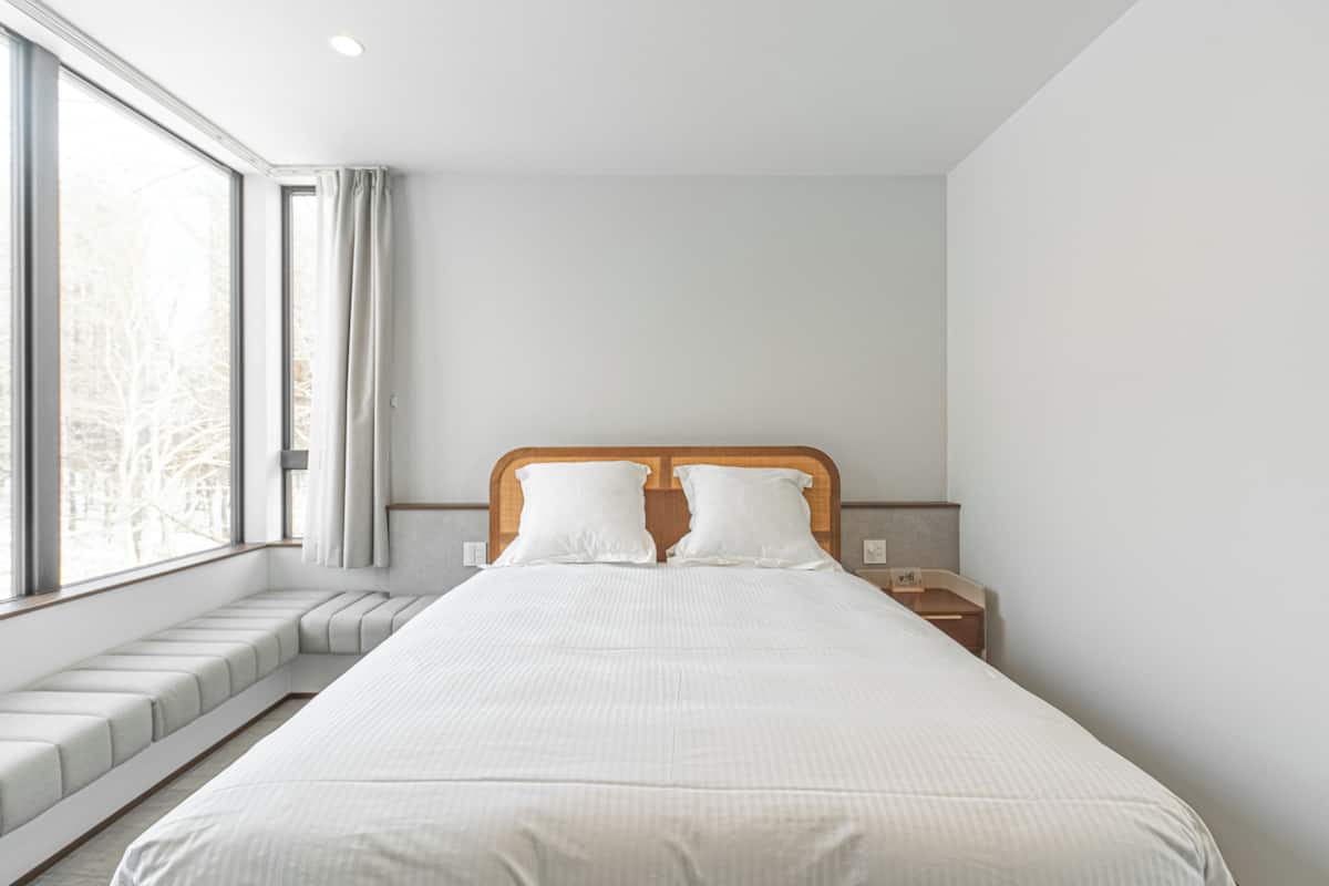 A modern hotel room with a large bed, a bedside table, a nightstand, a window with a view of trees, and a built-in bench along the window.