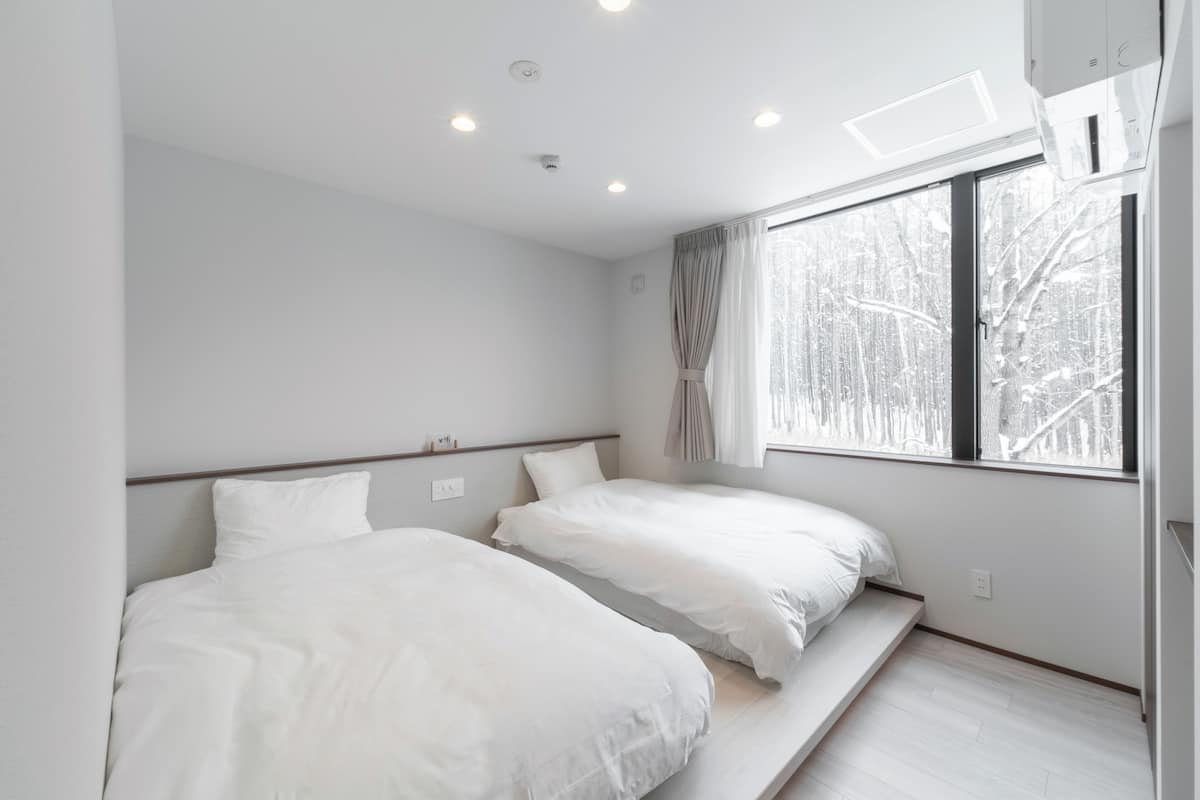 A modern hotel room with two beds, a large window with a view of trees, and a built-in bench.