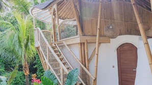 Una estructura de madera con una escalera que lleva a una puerta, rodeada de exuberante vegetación y un sendero de piedra.