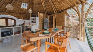 Un comedor rústico de madera con una gran mesa y sillas, una zona de cocina con electrodomésticos modernos y un techo de bambú.