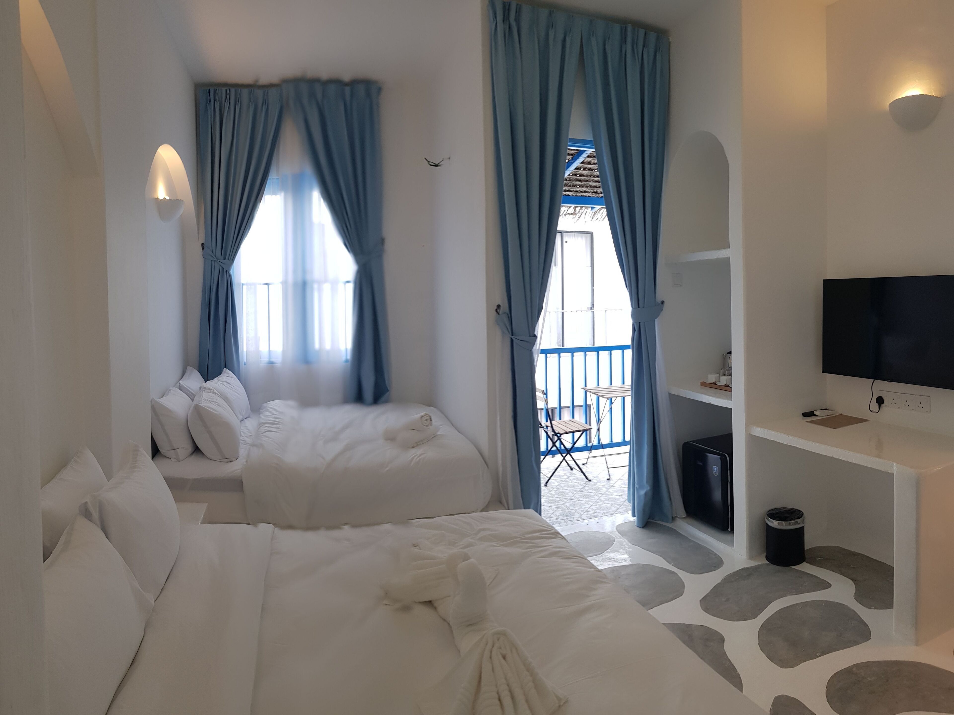 A hotel room with two beds, a balcony with blue curtains, a TV, and a small table.
