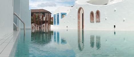 A clear swimming pool with a tiled edge, surrounded by white buildings with blue accents and arched windows.
