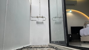 Un chemin dallé de pierres mène à un espace de douche clos par des parois de verre, où l’on peut voir une tuyauterie et un pommeau de douche.