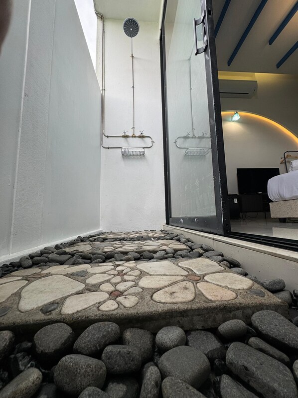 Un chemin dallé de pierres mène à un espace de douche clos par des parois de verre, où l’on peut voir une tuyauterie et un pommeau de douche.