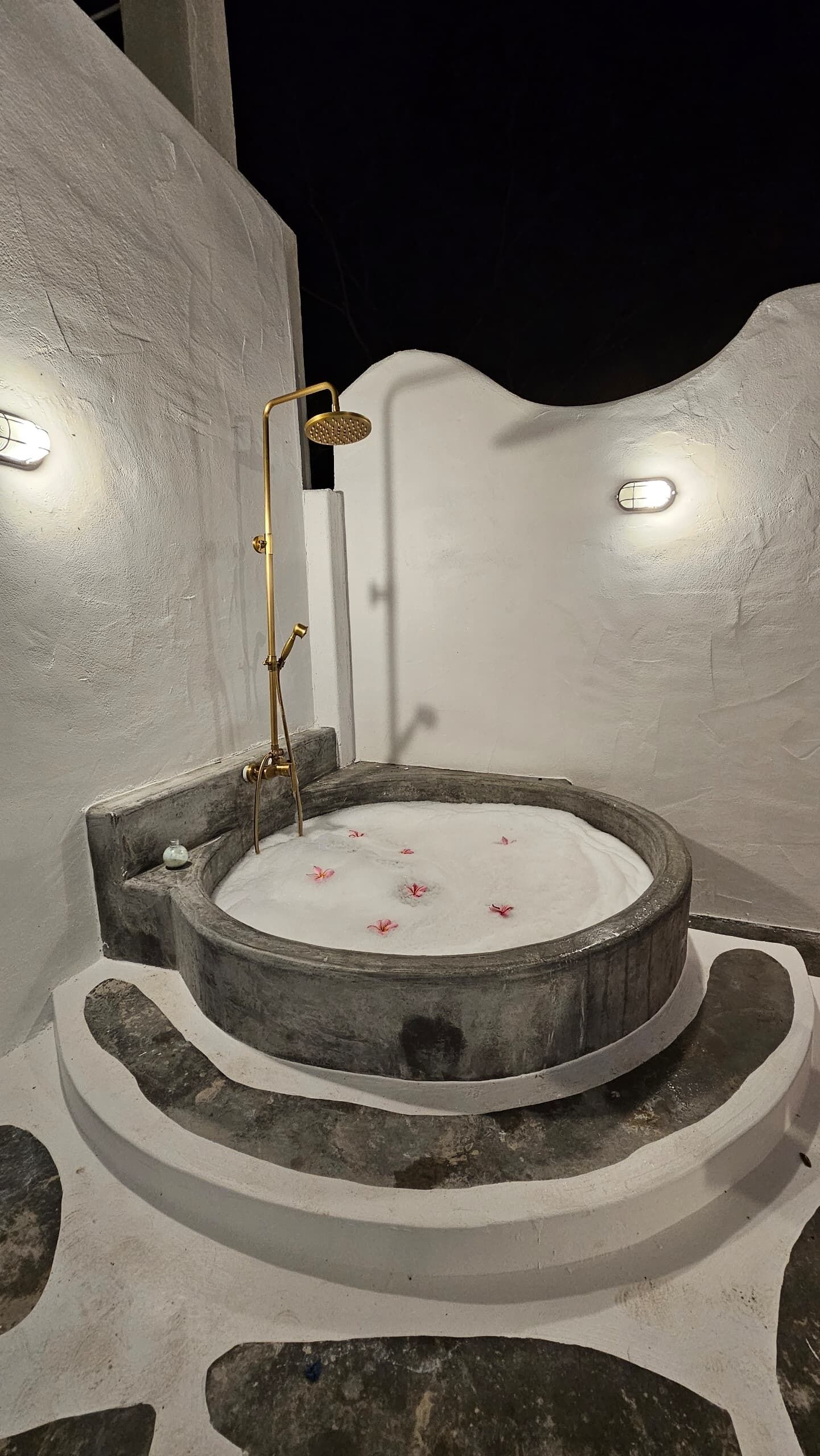 A circular hot tub with a showerhead, surrounded by stone steps and a white wall.