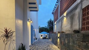Un chemin pavé de pierres mène à l’entrée d’un bâtiment, bordé de murs blancs et de portes bleues.