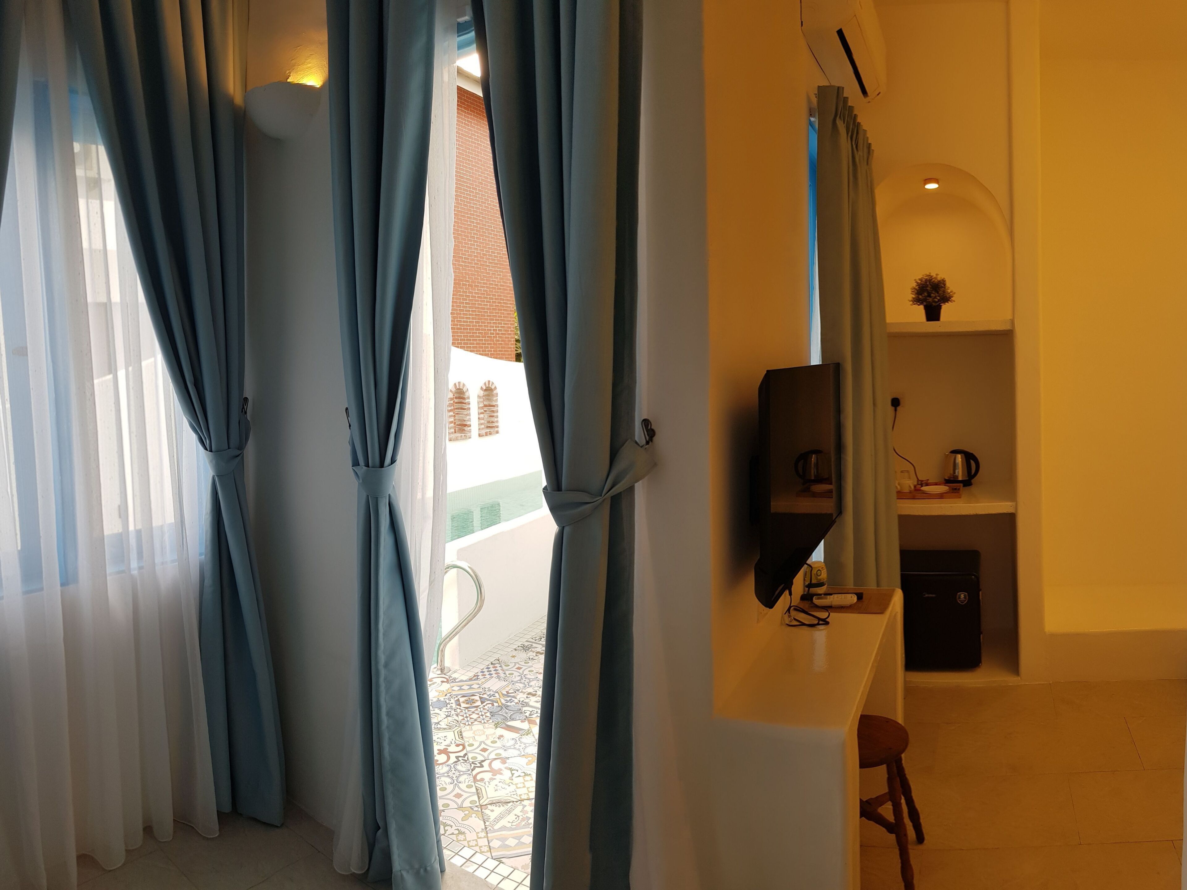 Una habitación con vistas a un edificio a través de cortinas, un televisor de pantalla plana montado en la pared, una pequeña zona de cocina con cafetera y suelo de baldosas.