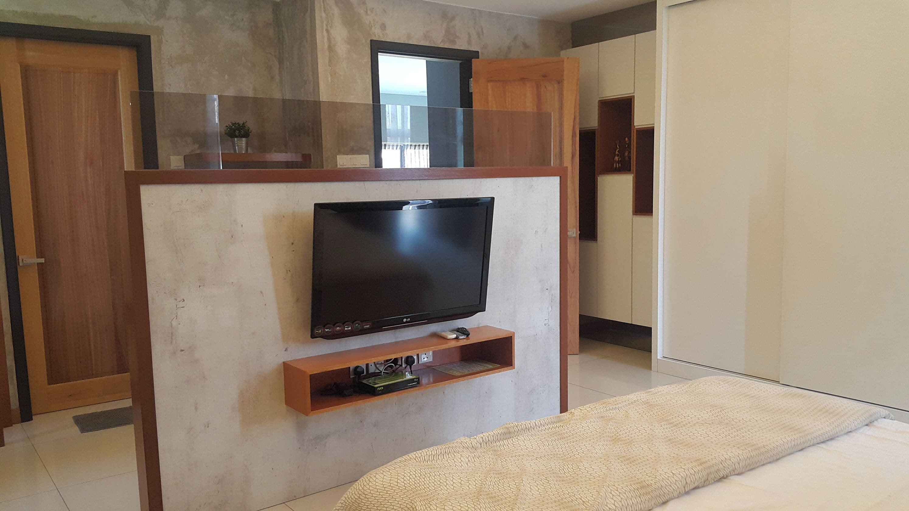 A bedroom with a TV mounted on a wooden wall, a bedside table with a remote, and a bed with a beige blanket.