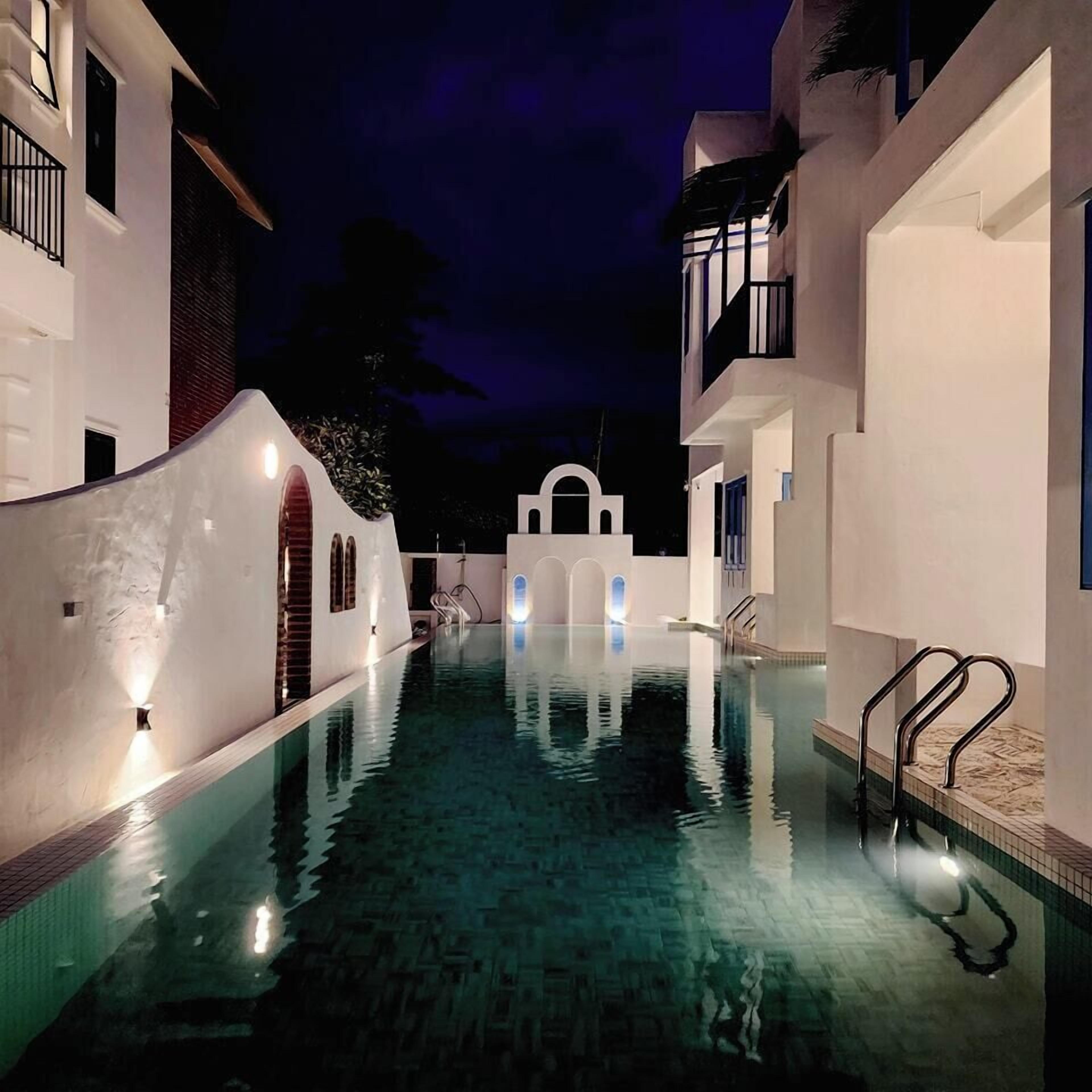 A swimming pool with a clear water surface, surrounded by white buildings with balconies and a central archway.