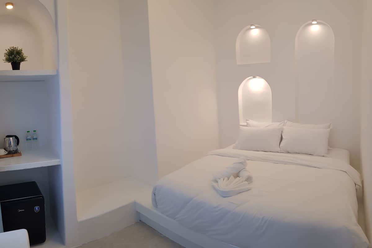 Ein minimalistisch eingerichtetes Hotelzimmer mit einem ordentlich bezogenen Bett, einem kleinen Nachttisch mit Wasserkocher und Tassen sowie einem Regal mit zwei Flaschen.