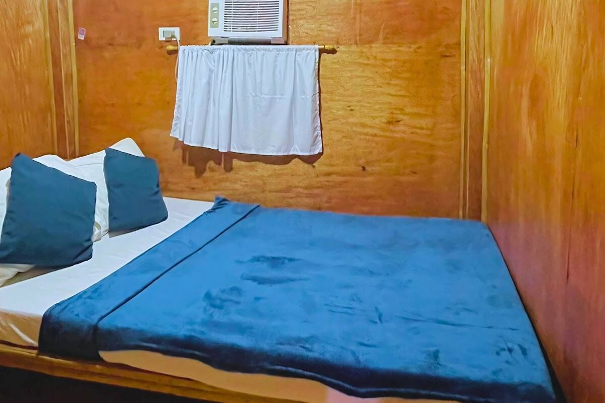 A single bed with a blue bedspread and two pillows, a white curtain hanging on a wooden wall, and a small air conditioning unit mounted above the curtain.