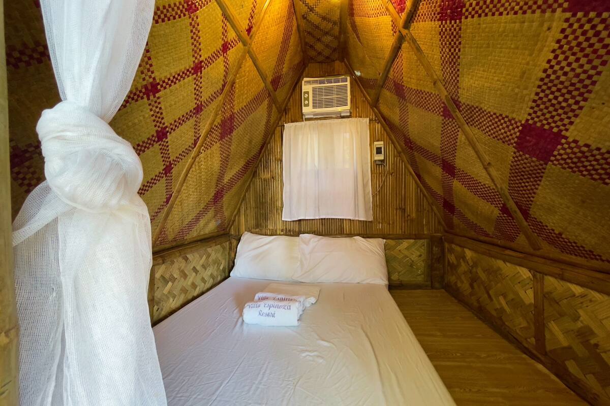 A cozy, triangular room with a bed, white linens, a curtain, and a small air conditioning unit.
