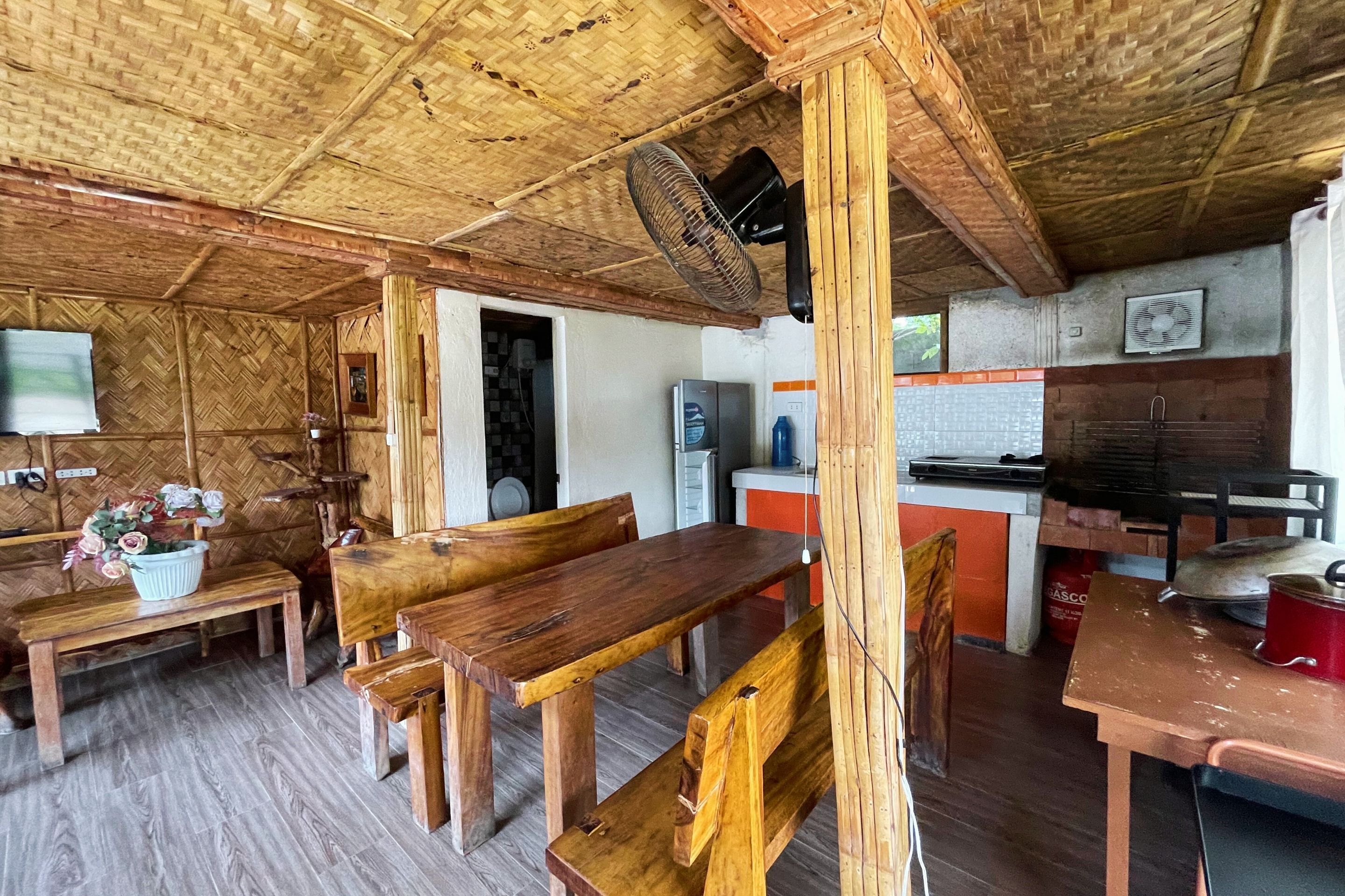 A rustic wooden interior with a herringbone-patterned ceiling, a wooden dining table with benches, a kitchen area with a refrigerator and stove, and a fan mounted on the ceiling.
